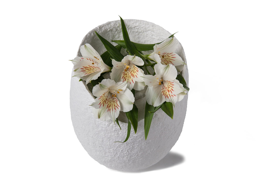 Picture of an ovoid biodegradable cremation urn made of white cotton fibre on sale at Muses Design Urns. View with flowers.
